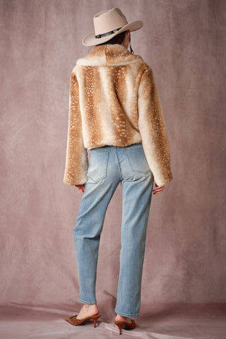 Person wearing a beige faux fur jacket and light blue jeans against a pinkish-beige background