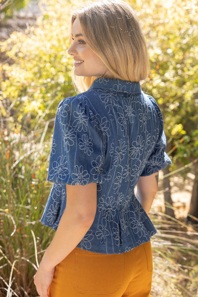 A woman wearing a blue short-sleeve blouse with a white floral pattern and orange pants, photographed outdoors in a sunlit garden setting.