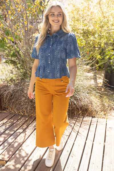 A woman stands on a wooden boardwalk outdoors wearing a blue short-sleeve button-up shirt with a white botanical pattern and bright mustard-yellow cropped pants, paired with white sneakers.