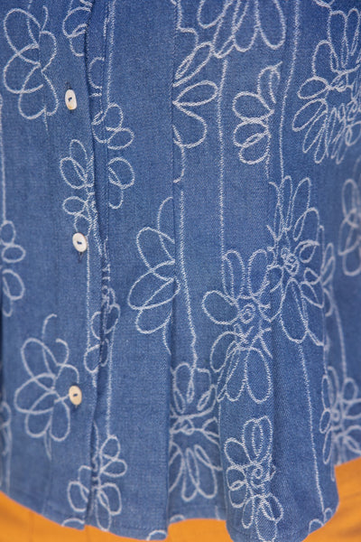 Close-up of a blue denim-style button-front garment with white floral line embroidery and small white buttons, shown against a small orange area at the bottom edge.