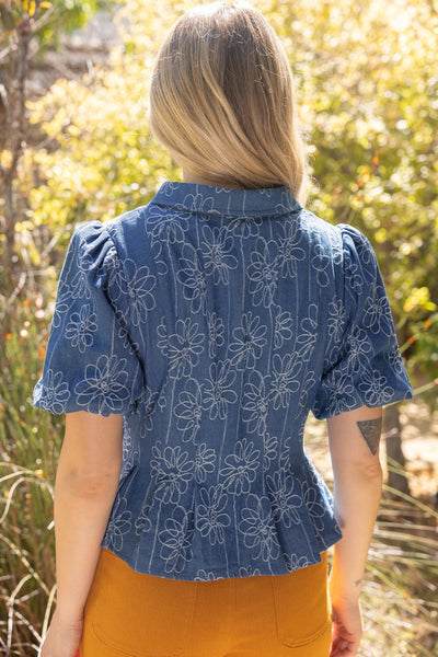 A woman viewed from behind wearing a blue short-sleeve blouse with a white floral pattern and a ruffled peplum hem, paired with mustard yellow pants, set against a blurred outdoor background of green foliage.