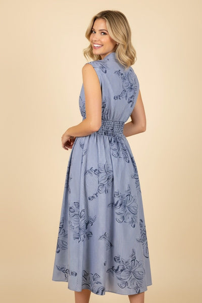 Woman wearing a blue floral pinstripe midi dress with a sleeveless design, smocked waist, front zipper, pockets, and A-line skirt