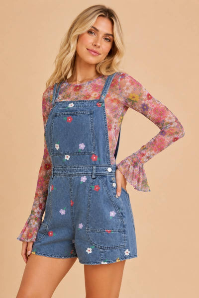 A woman modeling a pair of blue denim shorts with colorful embroidered flowers, worn over a long-sleeve floral print top, against a solid beige background.