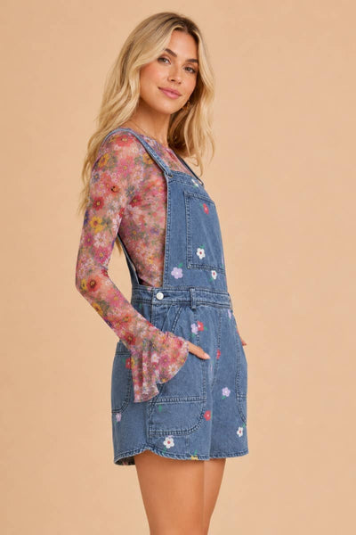 A woman modeling a blue denim overall dress with white floral embroidery, layered over a sheer long-sleeve floral mesh top, set against a solid beige background.