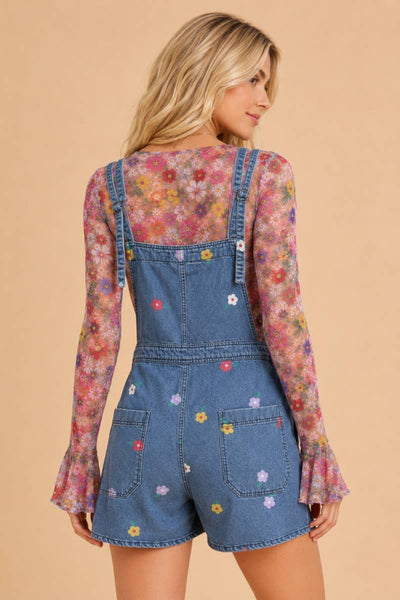 A woman shown from the back wearing a denim overall romper with long sleeves and a colorful floral print, set against a solid beige background.
