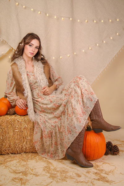 Woman in a floral dress and brown boots sitting on hay with pumpkins and string lights in the background