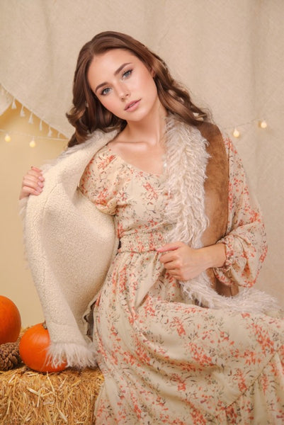 Woman in a floral dress holding a white shawl with pumpkins and hay bale in the background