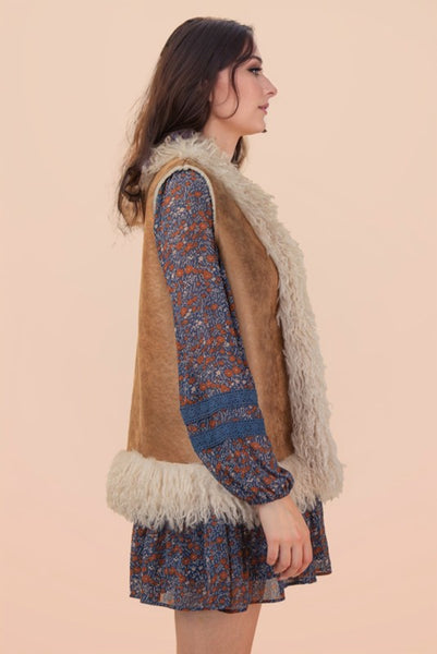 Woman wearing a patterned dress with a brown vest and fur trim against a beige background