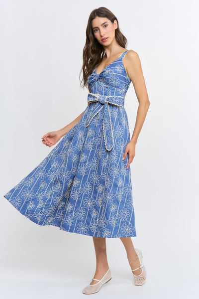 A woman modeling a sleeveless midi dress with a blue and white floral pattern, featuring a tie-waist belt and thin shoulder straps, set against a plain white studio background.