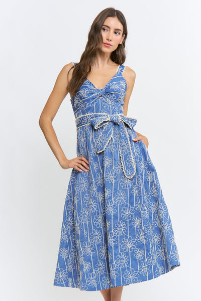 A woman modeling a sleeveless blue midi dress with a white floral and vertical stripe pattern, featuring a tie-waist belt and side pockets, set against a plain light gray background.
