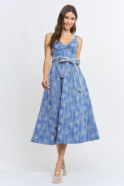 A woman modeling a sleeveless midi dress with a blue and white palm tree pattern, featuring a tie-waist belt and a V-neckline, set against a plain light gray studio background.