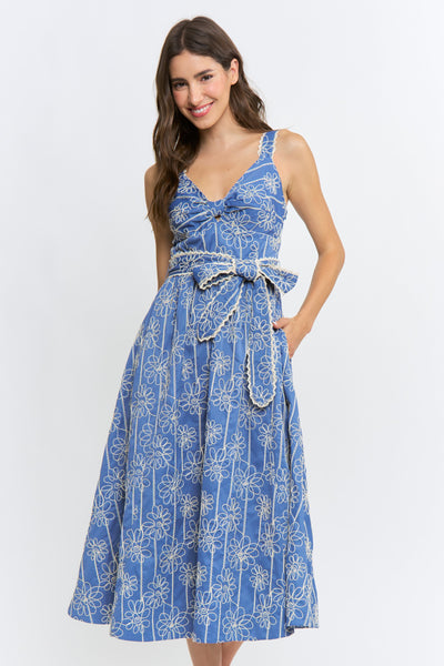 A woman modeling a sleeveless blue midi dress with a white floral and vertical stripe pattern, featuring a tie-waist belt and side pockets, set against a plain white studio background.