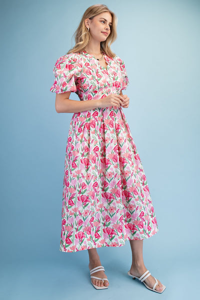 A woman modeling a short-sleeve midi dress with a pink and green floral print on a white background, paired with white strappy slide sandals, set against a solid light blue studio backdrop.