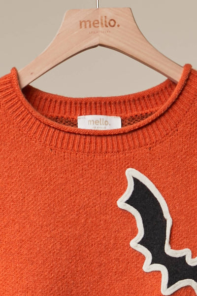 Close-up of a knit crewneck sweater in burnt orange hanging on a wooden hanger branded 'mello.' The sweater has a black bat applique with a white outline on the front and a visible 'mello.