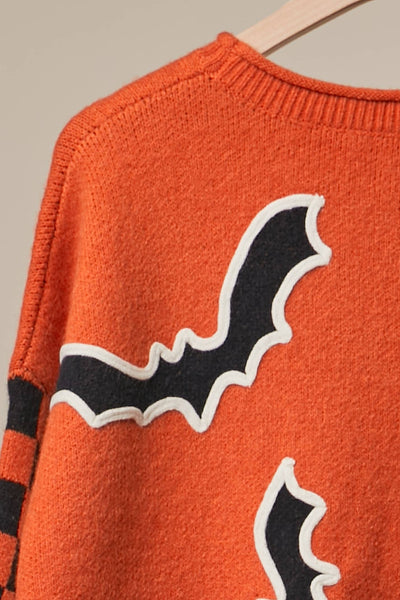 Close-up of an orange knit sweater with large black bat shapes outlined in white across the front, hanging on a wooden hanger against a light neutral background.