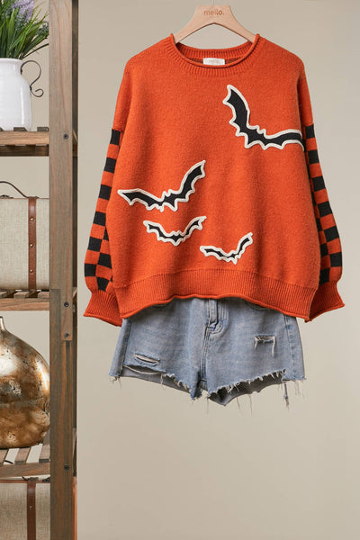 An orange crewneck sweater with black and white bat graphics and black-and-orange striped sleeves, styled over light-wash denim shorts on a wooden hanger against a beige wall.