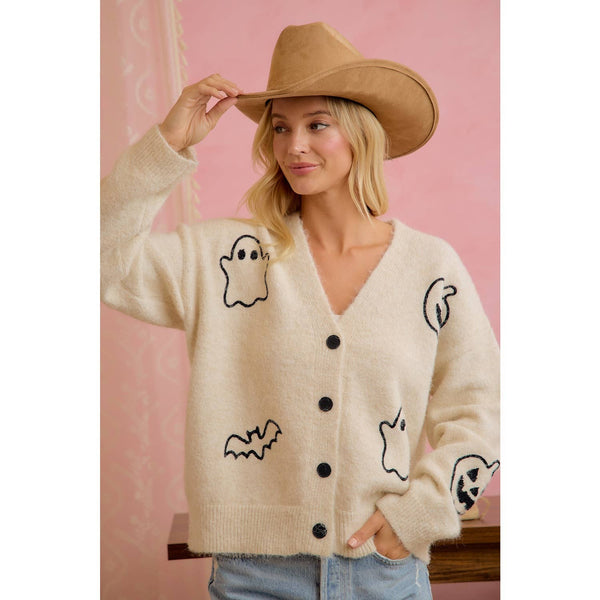 Woman wearing a cream-colored cardigan with black graphics, holding a brown cowboy hat against a pink background.