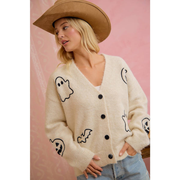 Woman wearing a cream cardigan with black ghost and bat designs, standing against a pink background.