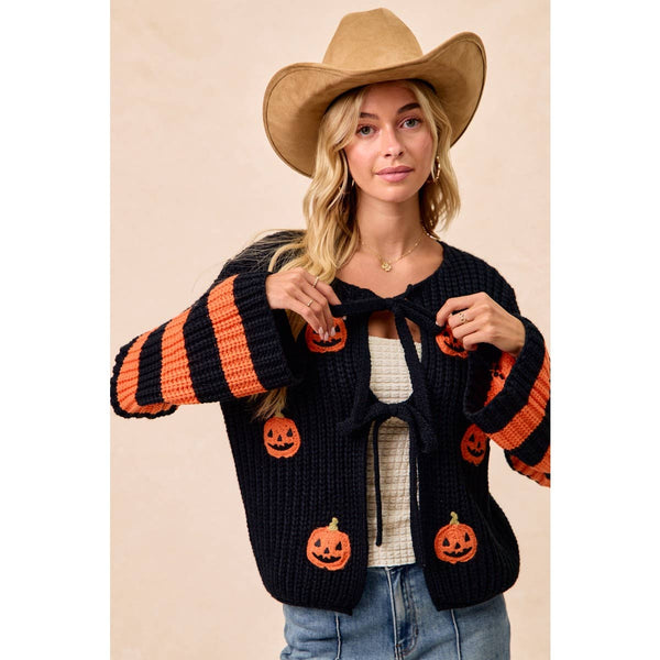A woman wearing a black knit cardigan with orange pumpkin patches and orange-and-black striped sleeves, styled with a tan cowboy hat and blue jeans against a plain beige background.