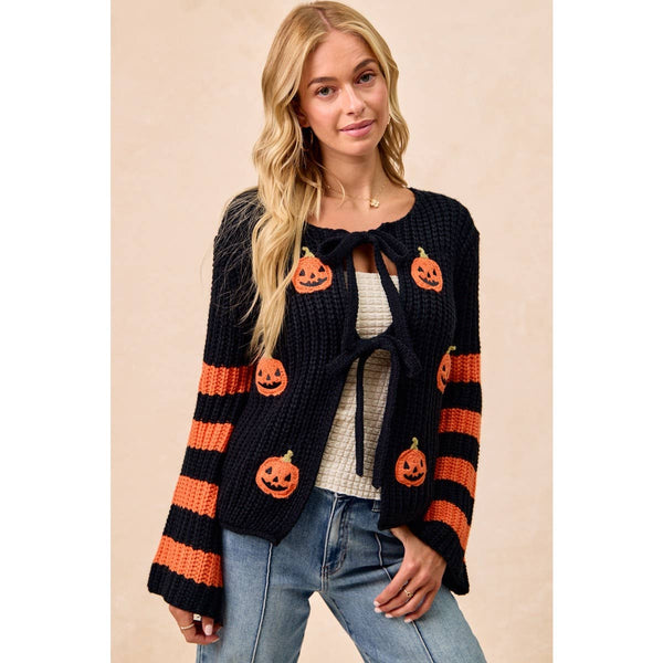 A woman modeling a black knit cardigan with orange pumpkin motifs and orange striped sleeves, worn over a cream top and blue jeans against a plain beige studio background.