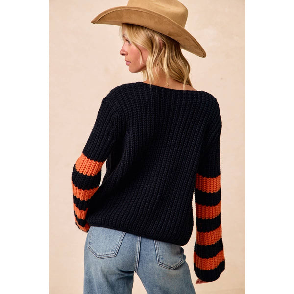 A woman wearing a black ribbed knit sweater with orange and black striped sleeves, paired with blue jeans and a tan cowboy hat, shown from the back against a plain beige background.