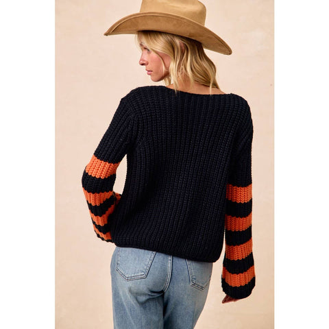 A woman shown from the back wearing a chunky knit sweater with a black body and orange and black striped sleeves, paired with light blue jeans and a tan cowboy hat against a beige studio background.