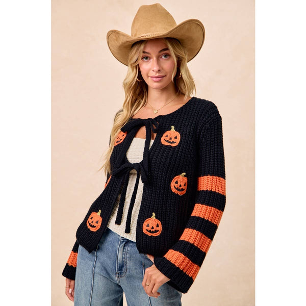 A woman wearing a black knit cardigan with orange pumpkin motifs and orange-and-black striped sleeves, styled with a tan cowboy hat, white top, and blue jeans against a plain beige studio background.