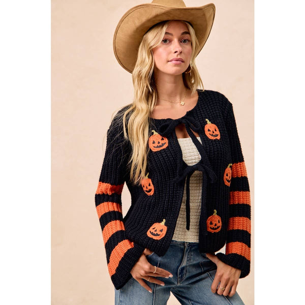 A woman wearing a black knit cardigan with orange pumpkin faces and orange-and-black striped sleeves, styled with a tan cowboy hat, white top, and blue jeans against a plain beige background.