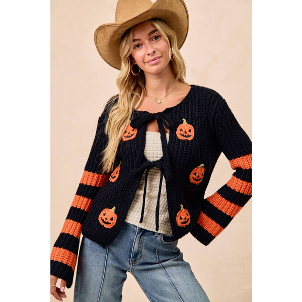 A woman wearing a black knit cardigan with orange pumpkin motifs and orange-and-black striped sleeves, styled with a tan cowboy hat, white top, and blue jeans against a plain beige background.