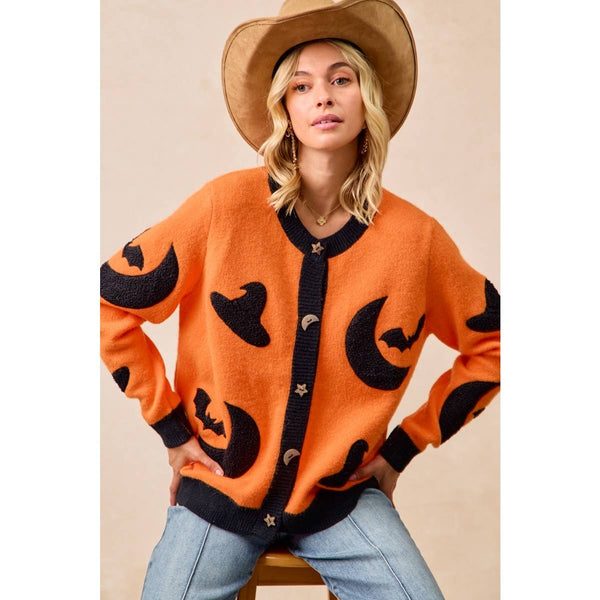 A woman wearing a bright orange knit cardigan with black moon and bat motifs, styled with a tan cowboy hat and light blue jeans against a beige studio background.