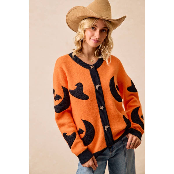 A woman wearing a bright orange knit cardigan with black moon and star motifs, paired with a tan cowboy hat and blue jeans, standing against a neutral beige background.