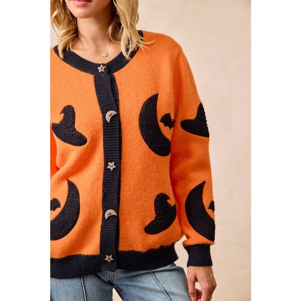 An orange knit cardigan sweater with black moon and witch hat motifs, featuring a black ribbed trim and star-shaped buttons, worn by a model.
