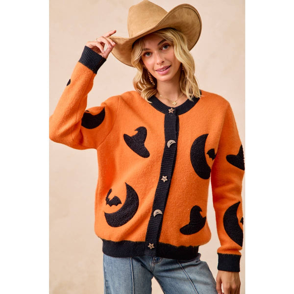A woman wearing a bright orange knit cardigan with black moon and bat motifs, a tan cowboy hat, and blue jeans, posed against a plain beige studio background.