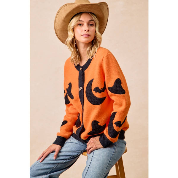 A woman wearing a bright orange knit cardigan with black bat and crescent moon motifs, styled with a tan cowboy hat and light blue jeans, seated against a neutral beige background.