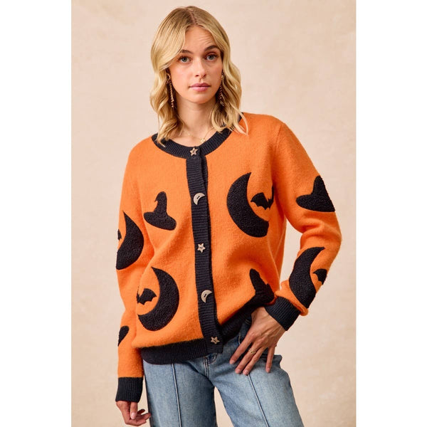 A woman wearing a bright orange knit cardigan with black moon, bat, and witch hat motifs, paired with light blue jeans, set against a warm beige studio background.
