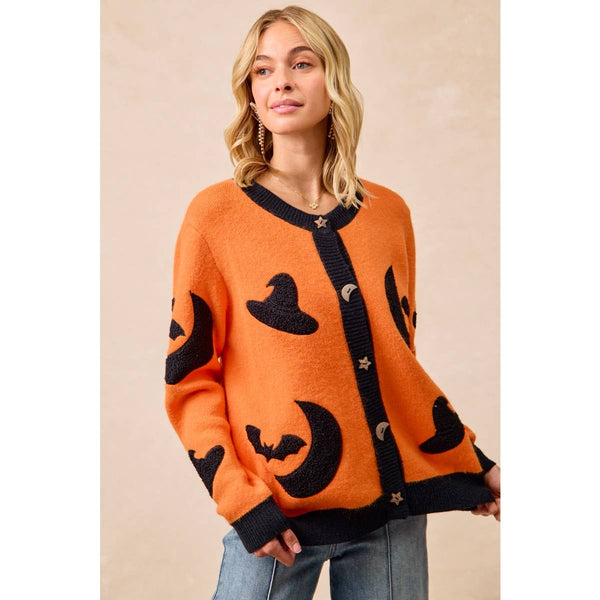 A woman wearing a bright orange knit cardigan with black trim and Halloween-themed motifs including witch hats, bats, crescent moons, and stars, paired with blue jeans against a neutral beige background.