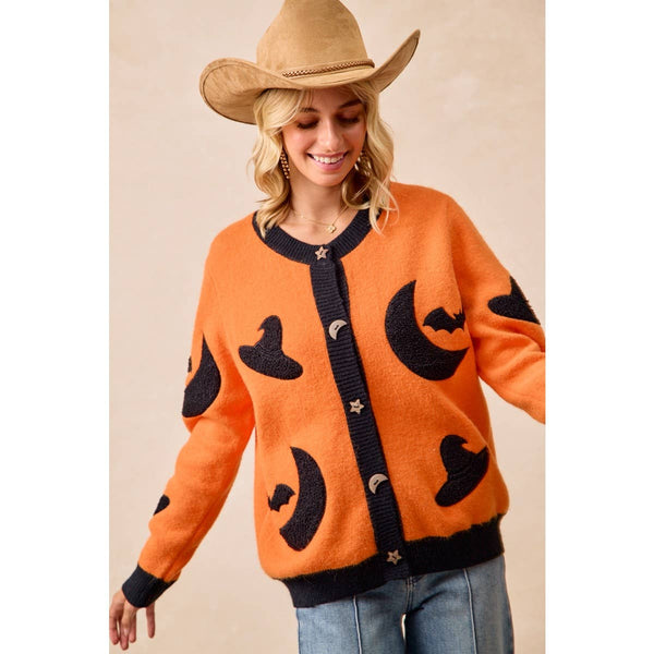 A woman wearing a fuzzy orange cardigan with black knit moon, bat, and witch hat motifs, paired with a tan cowboy hat and blue jeans, against a plain beige background.