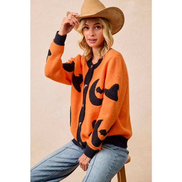 A woman wearing a bright orange knit cardigan with black abstract patches, blue jeans, and a tan cowboy hat, seated on a stool against a beige studio background.
