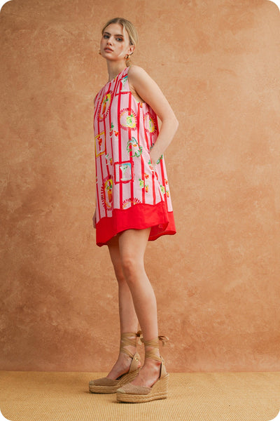 A woman modeling a sleeveless red and white striped dress with a colorful floral print, paired with beige wedge sandals, set against a textured warm brown backdrop.