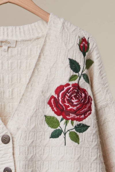An off-white textured knit cardigan sweater with a large red rose and green leaves embroidered on the chest, hanging on a wooden hanger against a neutral beige background.