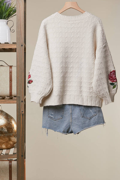 An off-white cable-knit sweater with red rose embroidery on the sleeves, hanging on a wooden hanger next to a wooden shelf, styled with light-wash denim shorts.