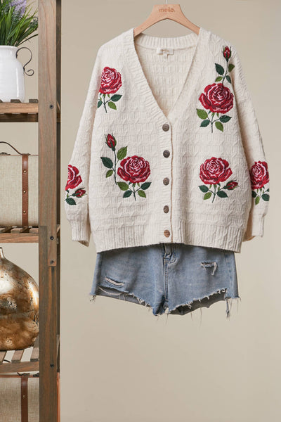 An off-white ribbed knit cardigan sweater with red rose embroidery, styled on a wooden hanger with distressed denim shorts against a beige wall.