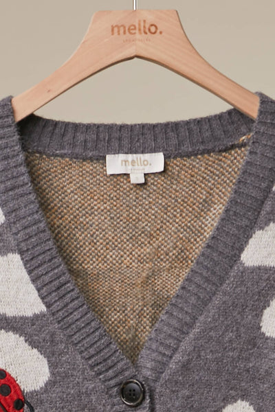 A close-up of a gray knit cardigan sweater with a brown and beige textured inner lining, hanging on a light wood hanger branded 'mello.' The cardigan has a V-neck, dark buttons, and a partial red-and-black polka dot pattern visible at the lower left.