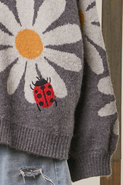 Close-up of a gray knit sweater featuring a large white daisy with a yellow center and a red ladybug motif, paired with light blue distressed jeans.