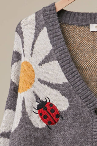 A close-up of a gray knit cardigan sweater featuring a large white daisy with a yellow center and a red ladybug motif on the front, hanging on a wooden hanger against a neutral beige background.