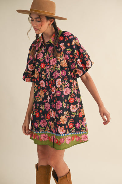 A woman wearing a short-sleeve black dress with a vibrant multicolor floral print, styled with a tan wide-brim hat and brown boots against a plain beige background.