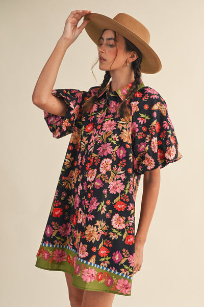 A woman wearing a short-sleeve black button-up dress with a vibrant multicolor floral print, styled with a wide-brimmed tan hat against a solid beige background.