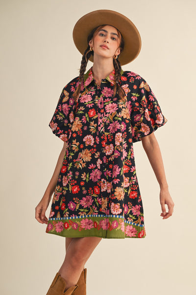 A woman wearing a short-sleeve button-up shirt dress with a vibrant multicolor floral print on a black background, styled with a wide-brimmed brown hat and brown boots against a solid beige studio background.