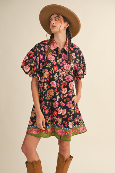 A woman modeling a short-sleeve black floral mini dress with puff sleeves, a pointed collar, and a green patterned hem, styled with a wide-brim tan hat and brown boots against a plain beige background.