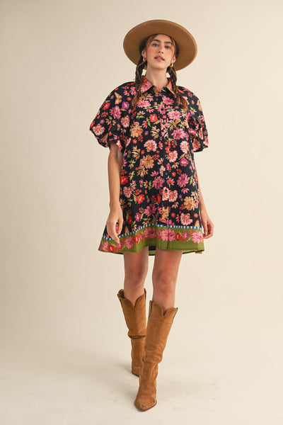 A woman modeling a short-sleeve black floral mini dress with a green patterned hem, styled with a tan wide-brim hat and tan knee-high boots against a plain beige studio background.